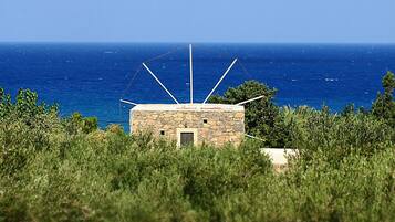 A stone building with a sloped roof, surrounded by greenery and overlooking a clear blue sea.