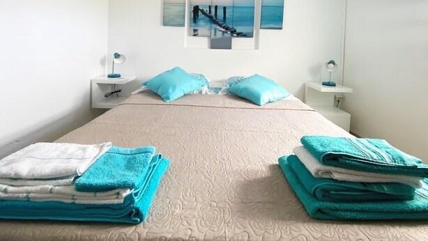 Una cama bien hecha con una colcha color crema, dos almohadas azules y una pila de toallas dobladas a cada lado. En la pared, hay una obra de arte que representa un muelle que se adentra en el mar.