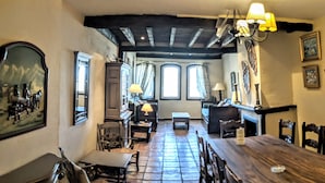 Un espace repas avec une longue table en bois, des chaises et un lustre. Il y a des fenêtres avec des rideaux et un tableau au mur.