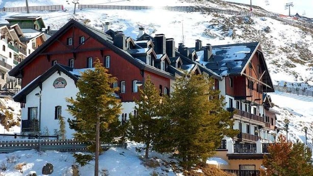 Un bâtiment de style chalet traditionnel, doté d’un toit à forte pente, qui ressemble probablement à un h ôtel ou une maison d’hôtes, entouré de montagnes enneigées et avec un télésiège visible au loin.