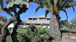 A stone building with a tiled roof, surrounded by a well-maintained garden with palm trees and cacti.