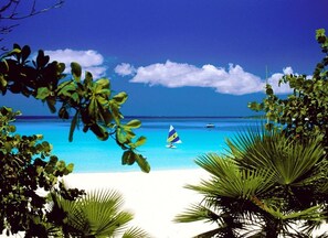 A beach with clear turquoise water, a sailboat, and lush greenery framing the view.