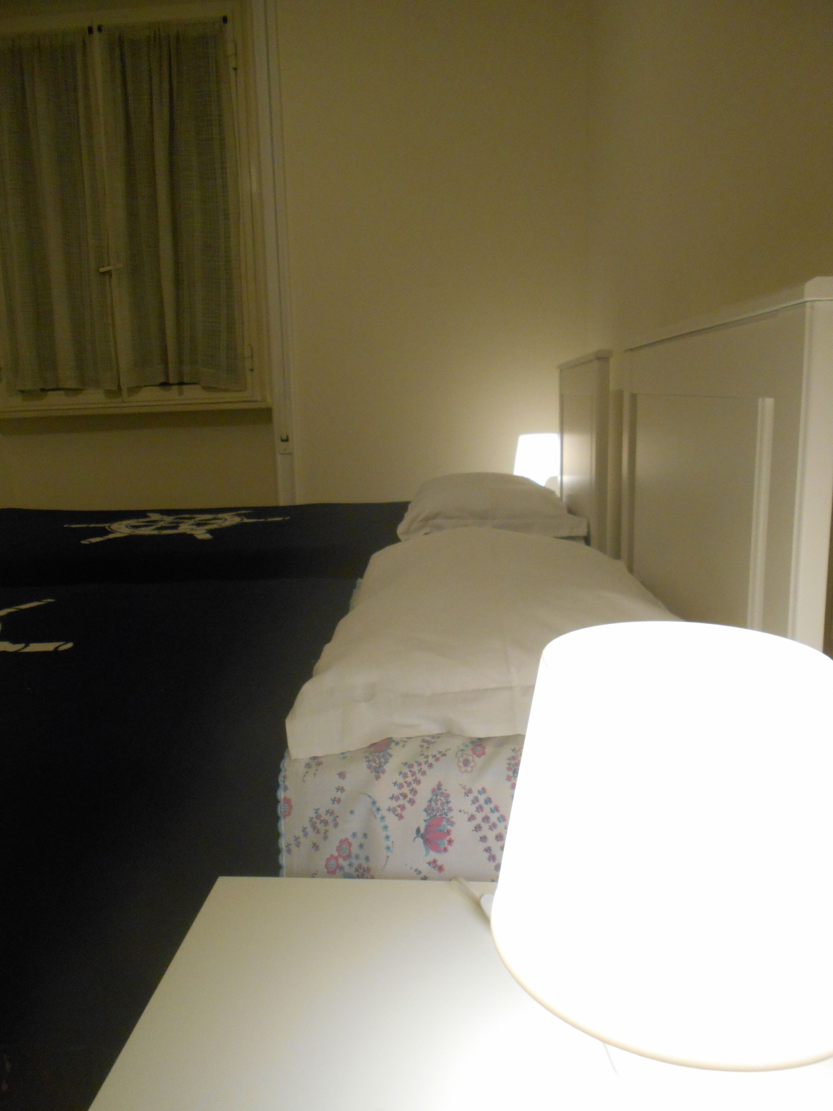 A single bed with a white headboard and a blanket, a bedside table, and a window with curtains.