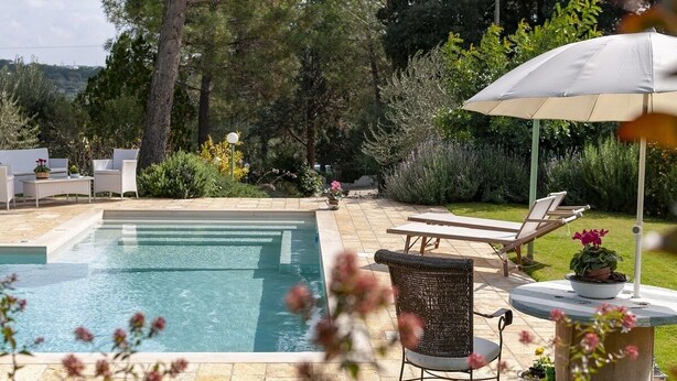 A swimming pool with lounge chairs and a table, surrounded by greenery and a white umbrella.