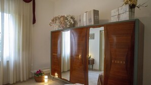 A bedroom with a wooden wardrobe, a bed with a floral bedspread, a bedside table with a lamp, and a window with curtains.