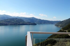 Vista panorámica de un lago rodeado de montañas, tomada desde un balcón con barandilla de vidrio.