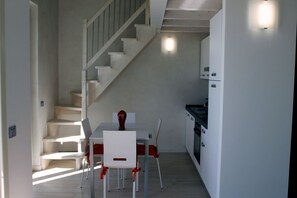 Un comedor con mesa y sillas blancas, una escalera y una zona de cocina.