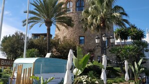 A historic stone building with arched windows and a balcony, surrounded by palm trees and outdoor seating.