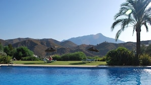 A swimming pool with lounge chairs and umbrellas, surrounded by greenery and mountains in the background.