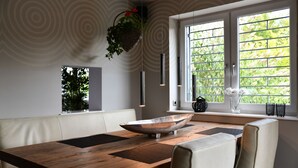 Un comedor moderno con una mesa de madera, asientos blancos acolchados, plantas colgantes y vistas a través de una ventana con celosía metálica.