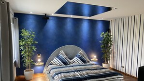 Un dormitorio moderno con una cama, dos plantas en macetas, una mesita de noche con una lámpara y una pared con rayas verticales.