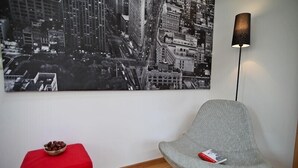 Een moderne kamer met een stadsgezicht schilderij, een rode poef, een grijze stoel en een vloerlamp.