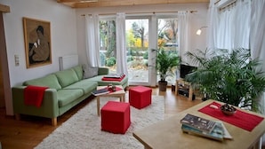 Een gezellige woonkamer met een groene bank, rode poefs, een houten salontafel en een groot raam met uitzicht op de omgeving.