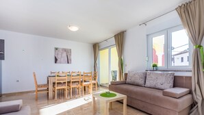 Ein Wohnzimmer mit einem beigefarbenen Sofa, einem Holzesstisch mit Stühlen, einem kleinen Holzhochentisch und einem großen Fenster mit Vorhängen.