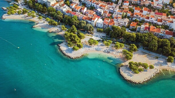 Eine Luftaufnahme einer Küstenstadt mit Strandabschnitt, kleinen Inseln und klarem türkisblauem Wasser, umgeben von Wohngebäuden mit roten Dächern.