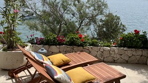 A terrace with wooden lounge chairs and a stone wall, overlooking a lake with boats and mountains in the background. - House directly by the sea (Klek)