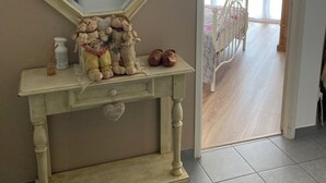 Una habitación con pared color crema, un espejo grande, una pequeña consola decorada y una cama al fondo.