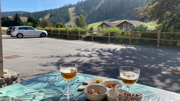 Una mesa con dos vasos de cerveza, platos de comida y una botella de salsa. Se observa un coche estacionado al fondo.