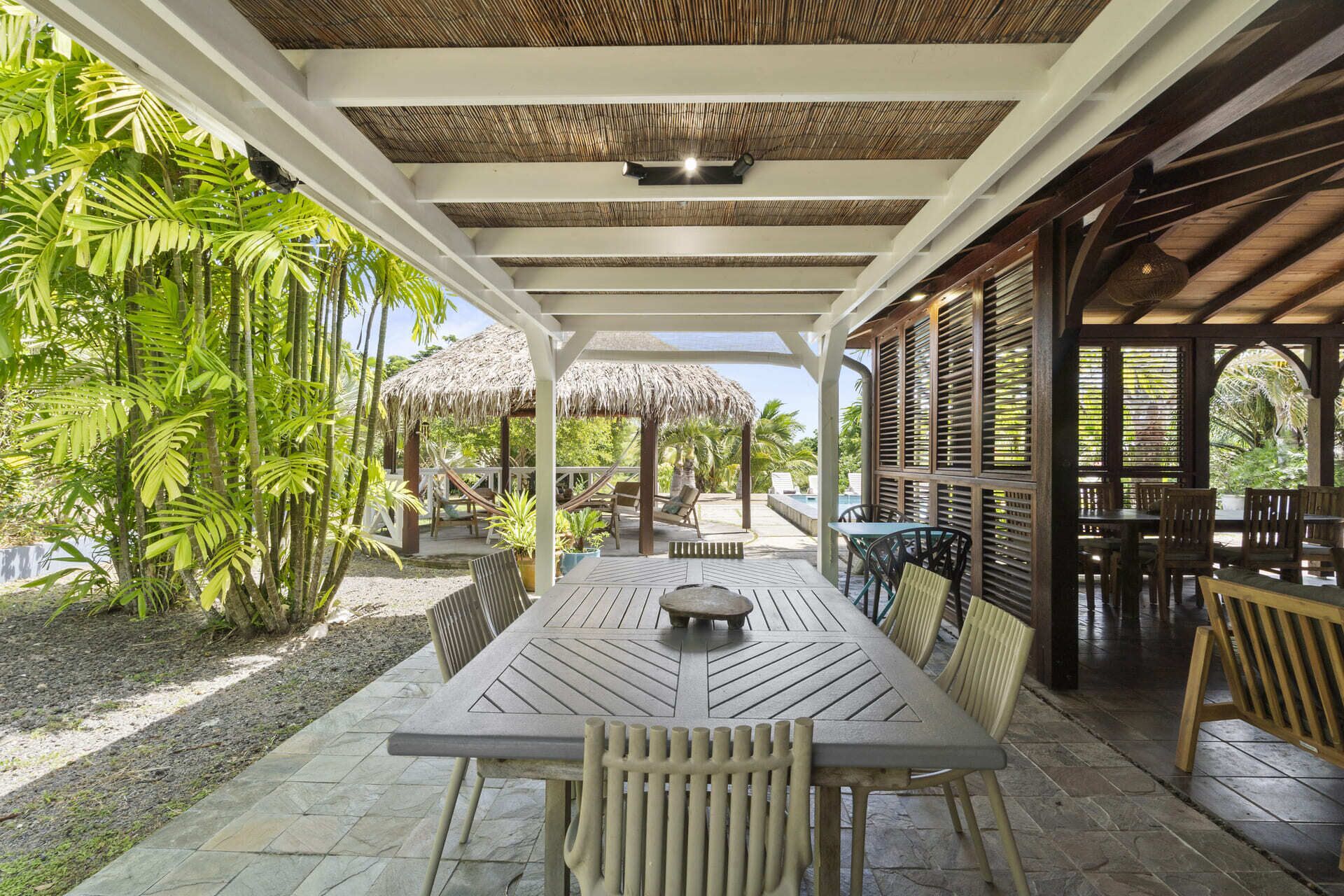 An outdoor dining area with a thatched-roof gazebo, surrounded by lush tropical plants.