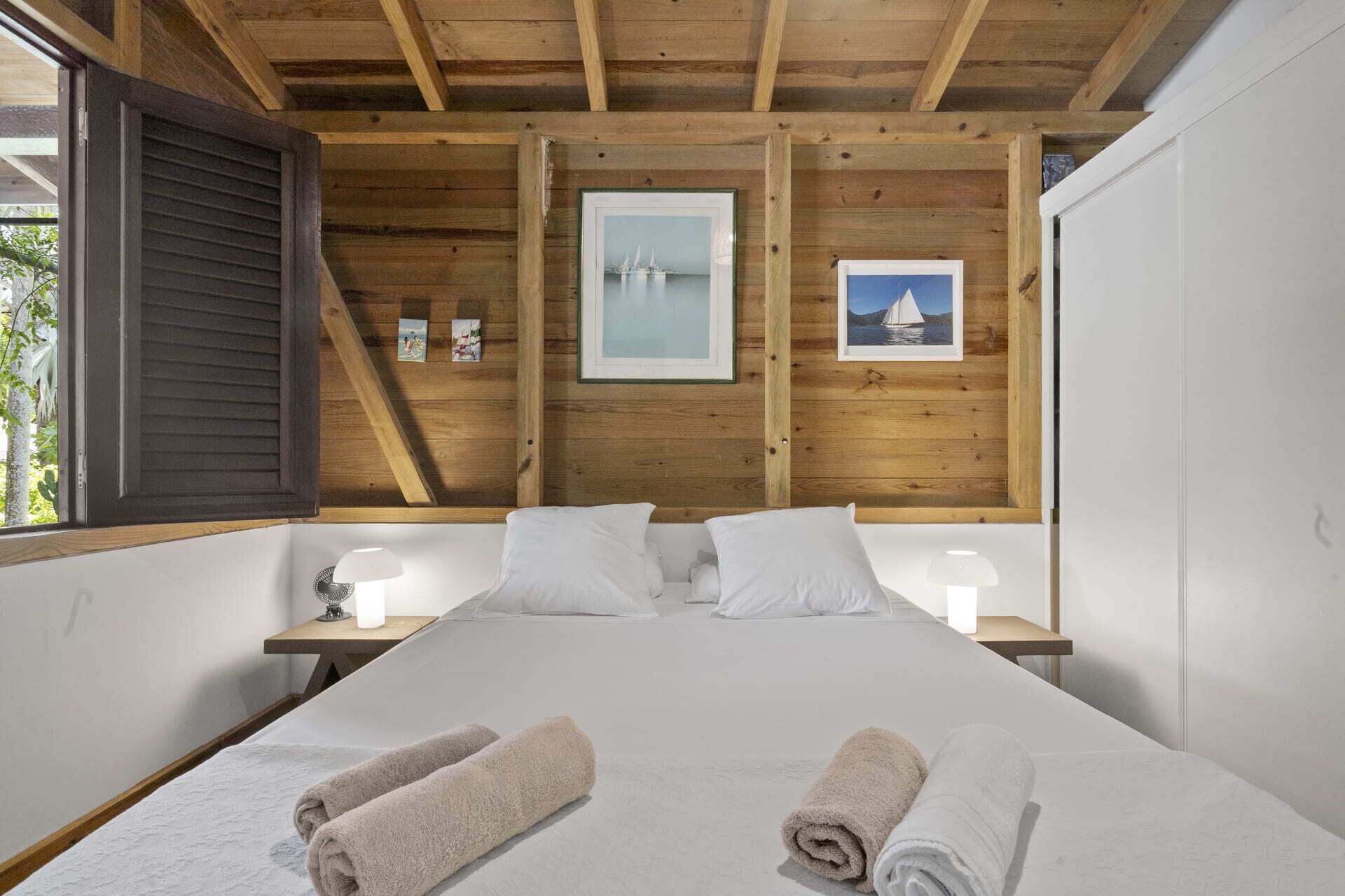 A bedroom with a wooden ceiling, a bed with white linens, two bedside tables with lamps, and framed pictures on the wall.