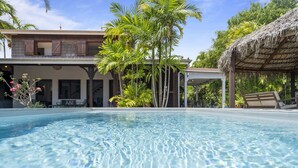 A clear swimming pool with a wooden deck area, surrounded by tropical vegetation and a thatched-roof structure. - Villa Colibri - Three Bedroom Villa, Sleeps 6 (Le Vauclin)