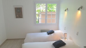 A room with two beds, a window, a wall-mounted light, and a small table with a lamp.