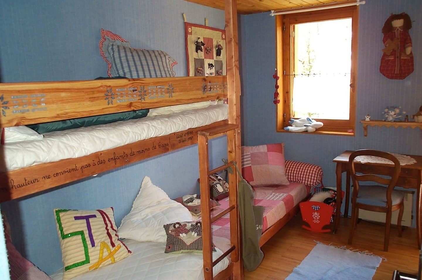 A dormitory room with bunk beds, a small desk, a chair, and a window with a view.
