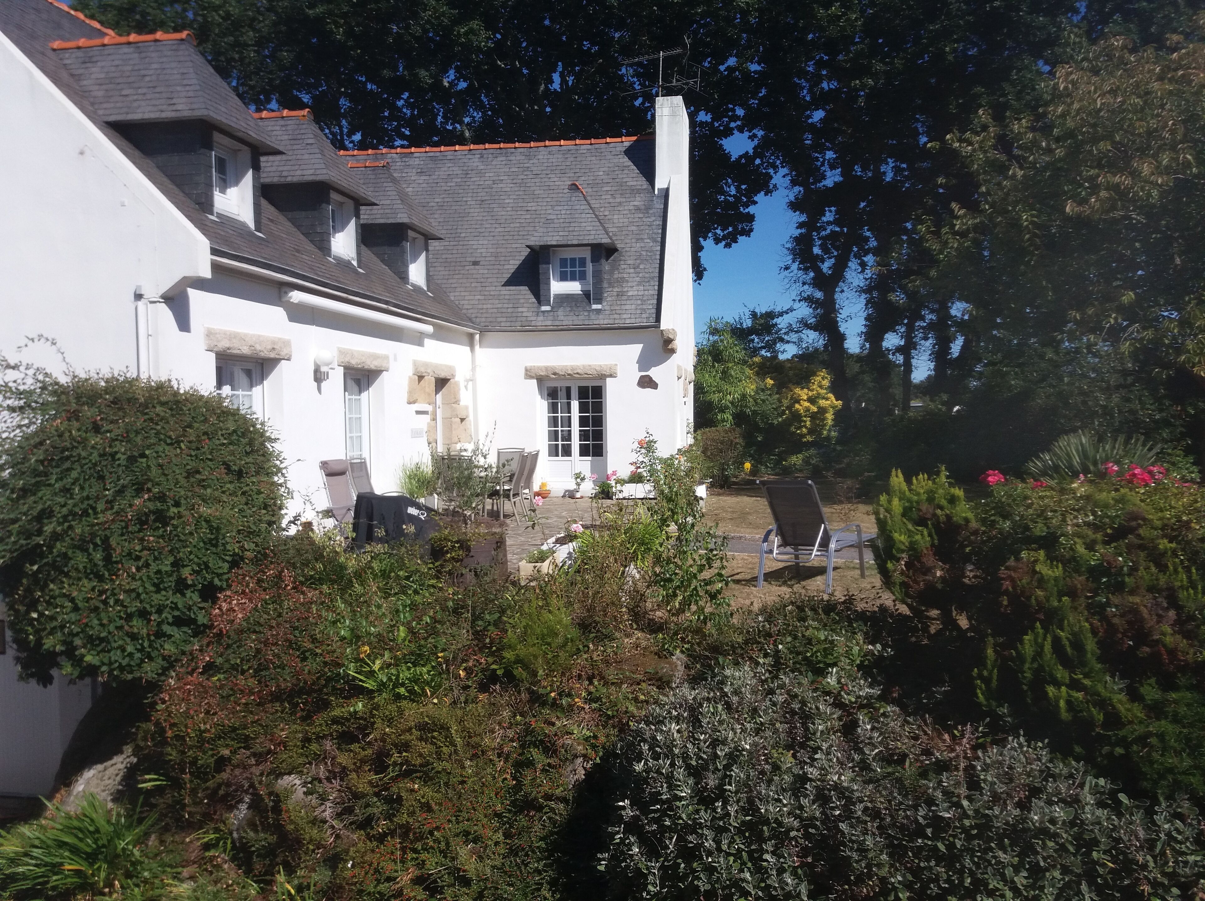Une maison à deux étages, dotée d’un toit en pente, d’une cheminée et d’un petit balcon. On y trouve des espaces de détente en plein air avec des chaises et une table.
