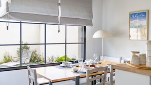Un coin repas avec une table dressée pour quatre personnes, de grandes fenêtres donnant sur un paysage verdoyant et un tableau encadré accroché au mur.