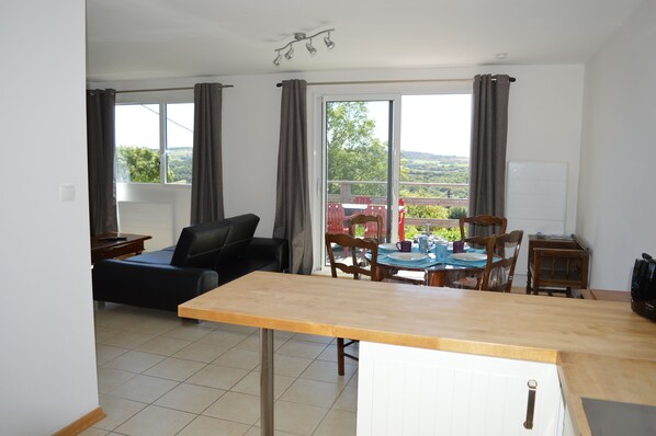 Un vaste espace de vie comprenant un canapé, une table à manger et des chaises. Une grande fenêtre, agrémentée de rideaux, offre une vue sur la verdure environnante, et un luminaire est fixé au plafond.