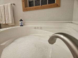 A bathroom with a bathtub filled with bubbles, a wooden-framed window, and a bottle of liquid soap. - Big Sky Cabin! Hot Tub! Fire Pit! Pool Table!   Private! Can hold Large Groups
 (Alto Pass)
