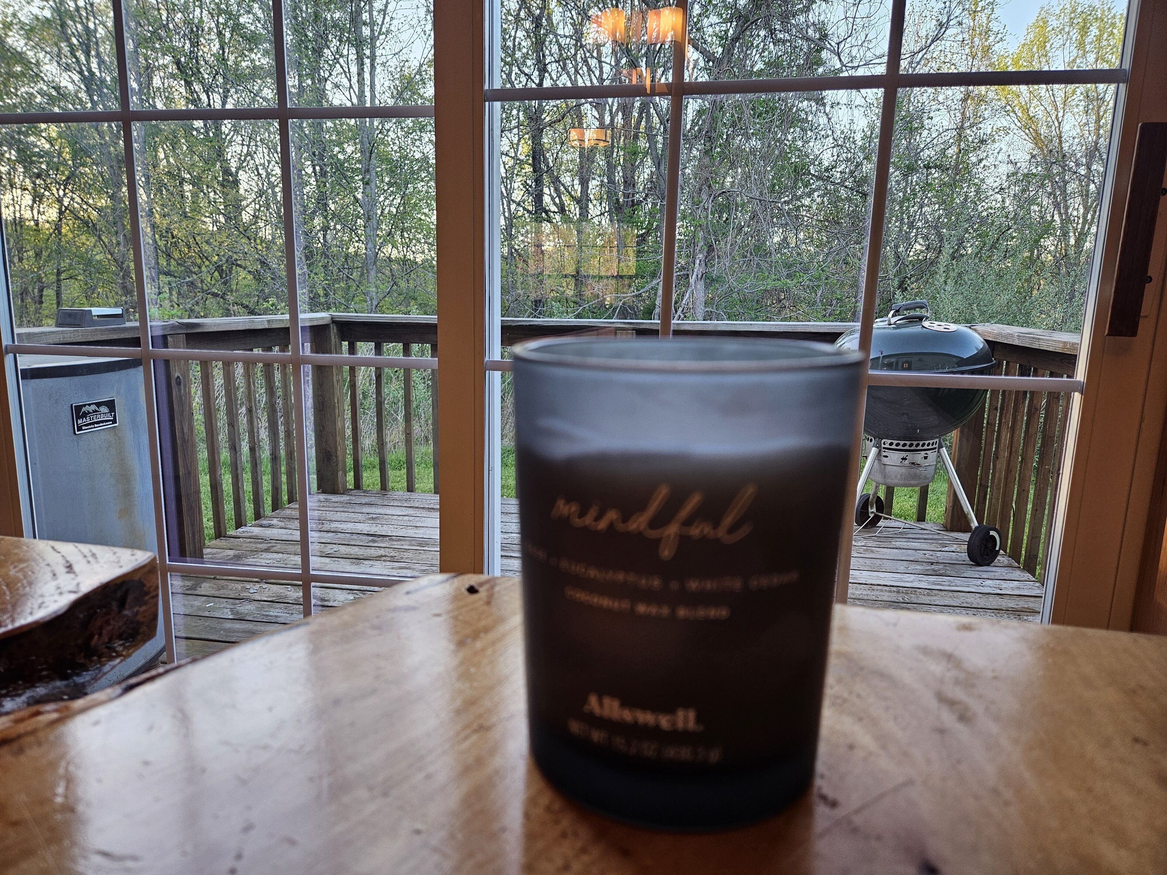 A candle on a wooden table with a view of a deck and a grill in the background.