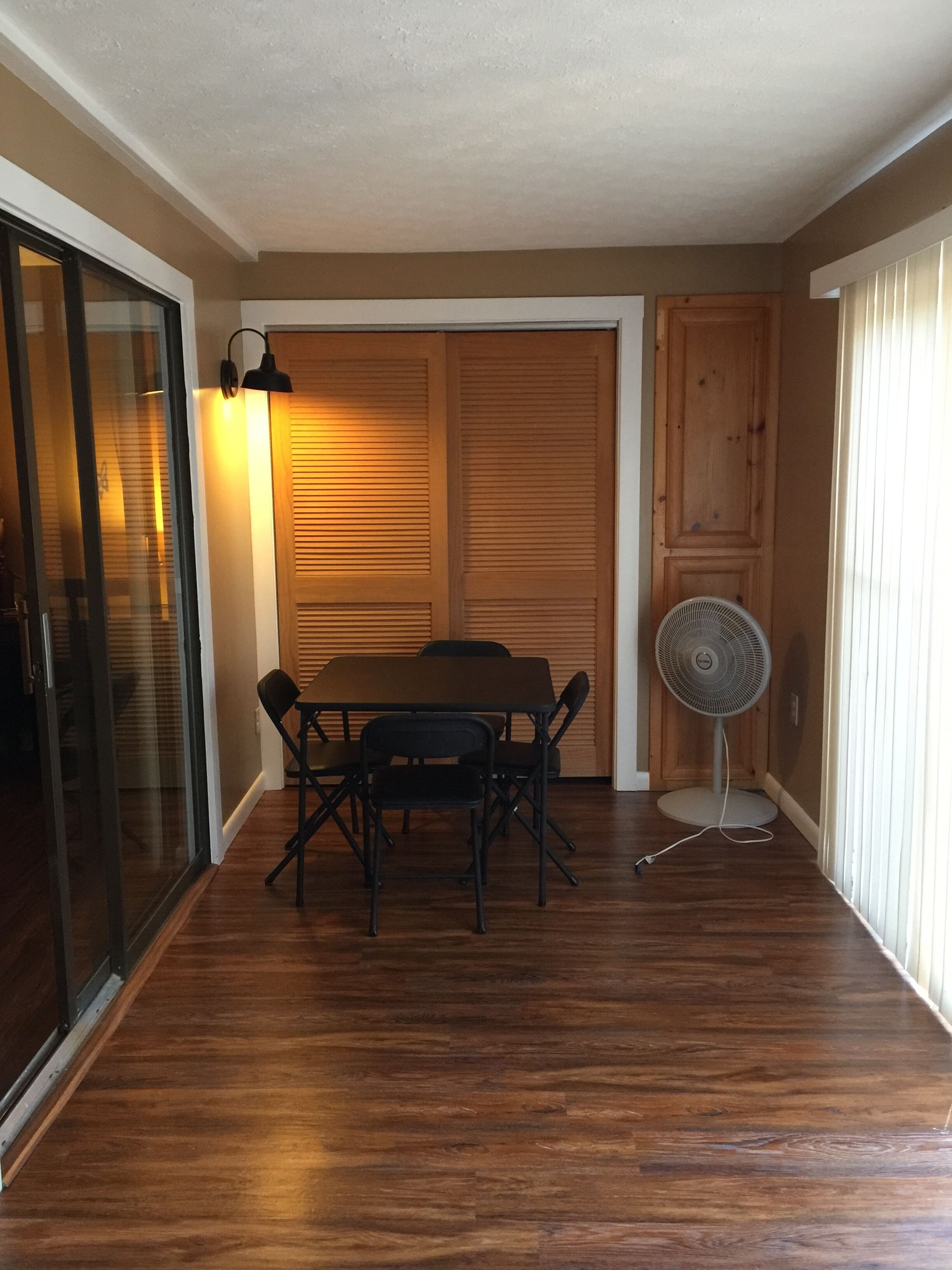 Quarto com piso de madeira, uma mesa com cadeiras, um ventilador e uma porta deslizante com persianas.