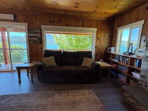 A cabin interior with a wooden ceiling, a couch with cushions, a bookshelf, and a view of a lake through a window.