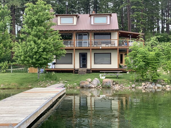 A two-story house with a dock extending into a calm lake, surrounded by trees. - 3 Bedroom Home On Waitts Lake With 150 Feet Of Lake Front (Valley)