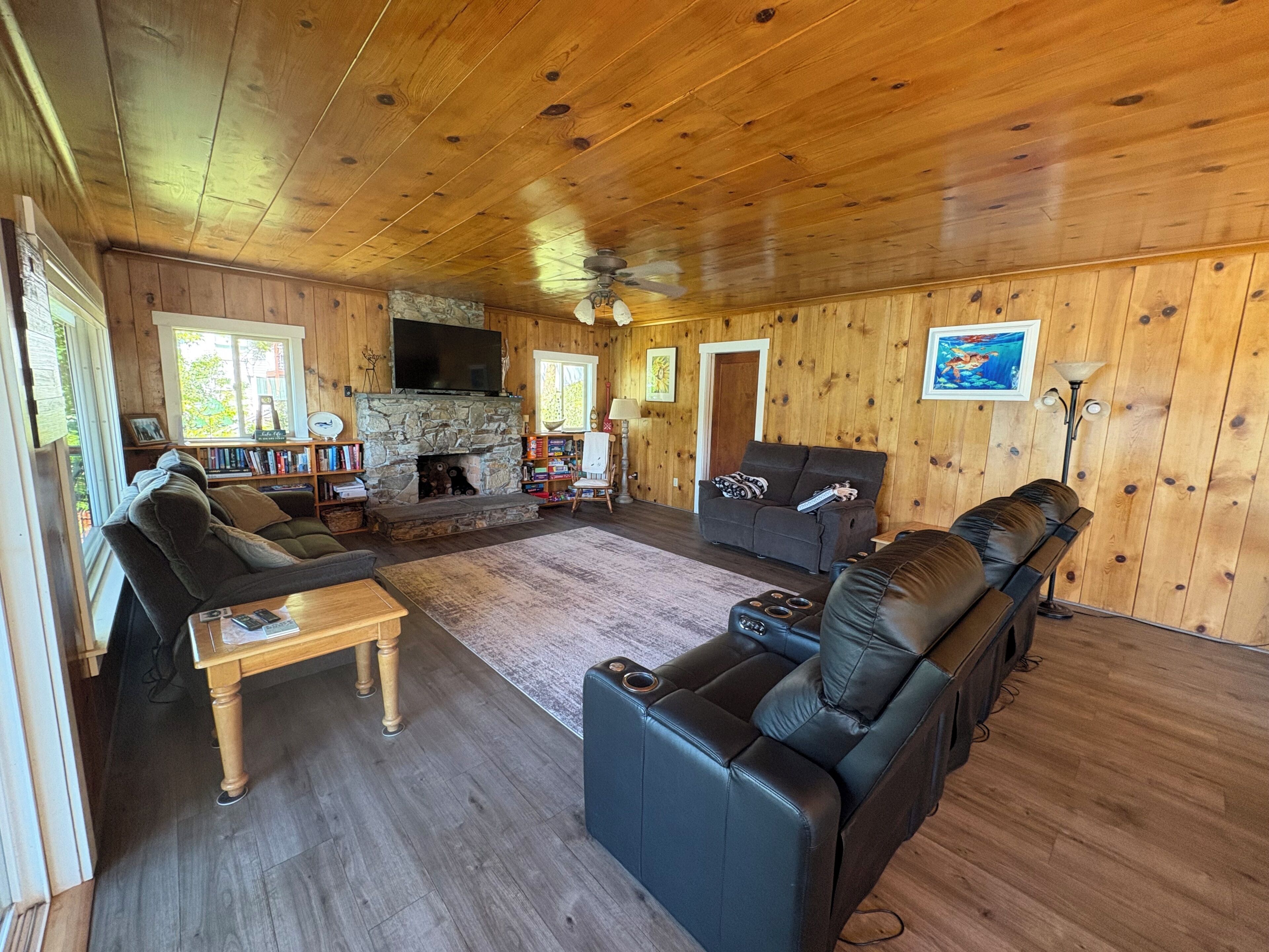 A log cabin interior with a stone fireplace, leather sofas, a wooden coffee table, and a television mounted on the wall.