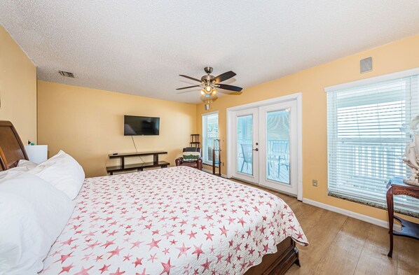 A bedroom with a bed, a ceiling fan, a TV, a window with blinds, and a door.