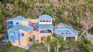 Uma casa grande e com vários andares, com piscina, cercada por vegetação exuberante.