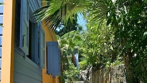 Uma trilha de pedra estreita ladeada por vegetação exuberante e um edifício amarelo com persianas azuis.