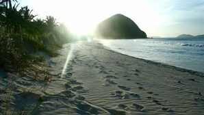 Uma praia com pegadas na areia, uma grande formação rochosa e o oceano ao fundo.
