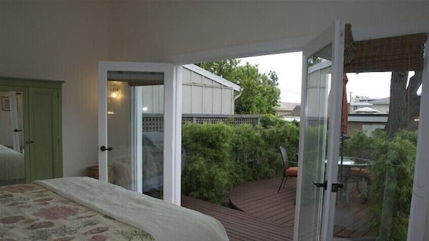 Ein Schlafzimmer mit einem großen Bett, einer Schiebetür zu einer Holzterrasse mit Tisch und Stühlen sowie Blick auf einen Garten und Nachbarhäuser.