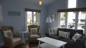 Ein Wohnzimmer mit einer Couch, zwei Sesseln, einem Couchtisch und einer Lampe. Es gibt drei Fenster und eine Tür.