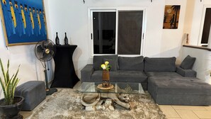 A modern living room with a grey sectional sofa, a glass coffee table, and a decorative sculpture. There is a large window with a view of the outside, a black floor lamp, and a painting on the wall.