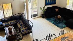 A spacious living area with a high ceiling, a balcony with yellow pillars, a black sofa, a patterned rug, and a tiled floor.