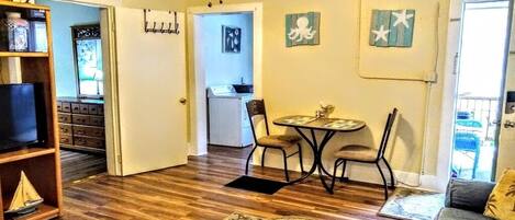 A living room with a wooden floor, a sofa, a coffee table, and a TV on a shelf. There is a dining area with a table and chairs, and a kitchen in the background.