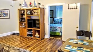 Een knus woongedeelte met een houten boekenkast, een televisie, een eettafel met decoratieve tafelonderleggers met een strandthema en een bed met een kleurrijk sprei.