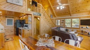 A wooden cabin interior with a kitchen, dining area, and a staircase leading to a loft.