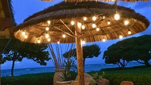 An outdoor seating area with a thatched umbrella and string lights, overlooking a body of water. - Gorgeous 1BR/2BA - Great Views & Reviews (Kapaa)