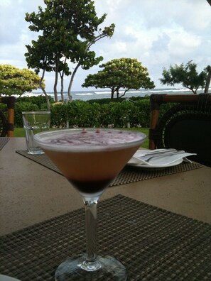 A martini glass with a layered drink on a table with a view of the ocean and trees in the background.