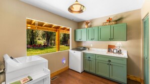 A kitchen with wooden flooring, a washing machine, a window with a view of a garden, and green cabinets.
